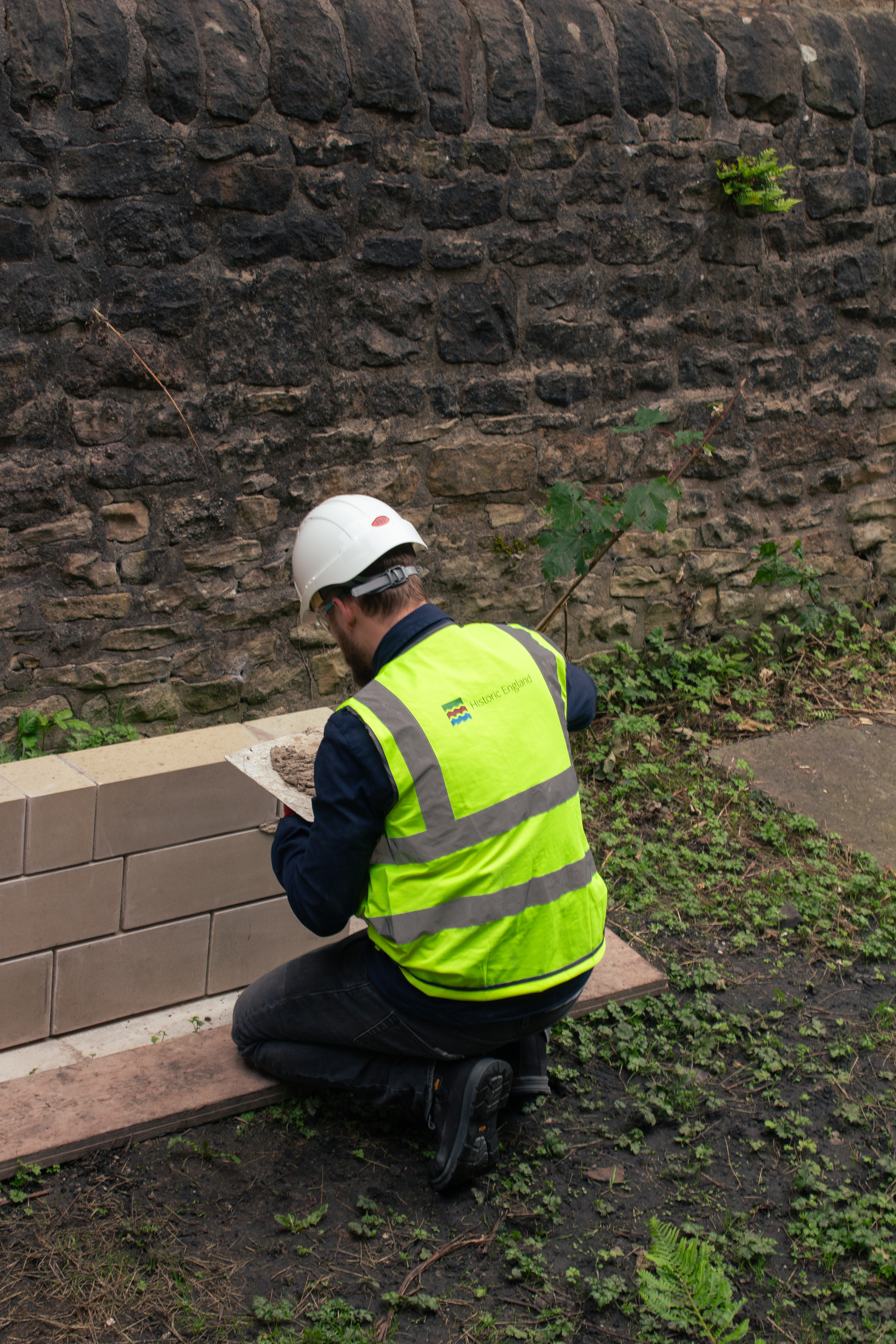 Image of trainee with Historic England hi-vis vest and white hard hat kneeling in front of a low practice wall practicing the application of lime mortar.