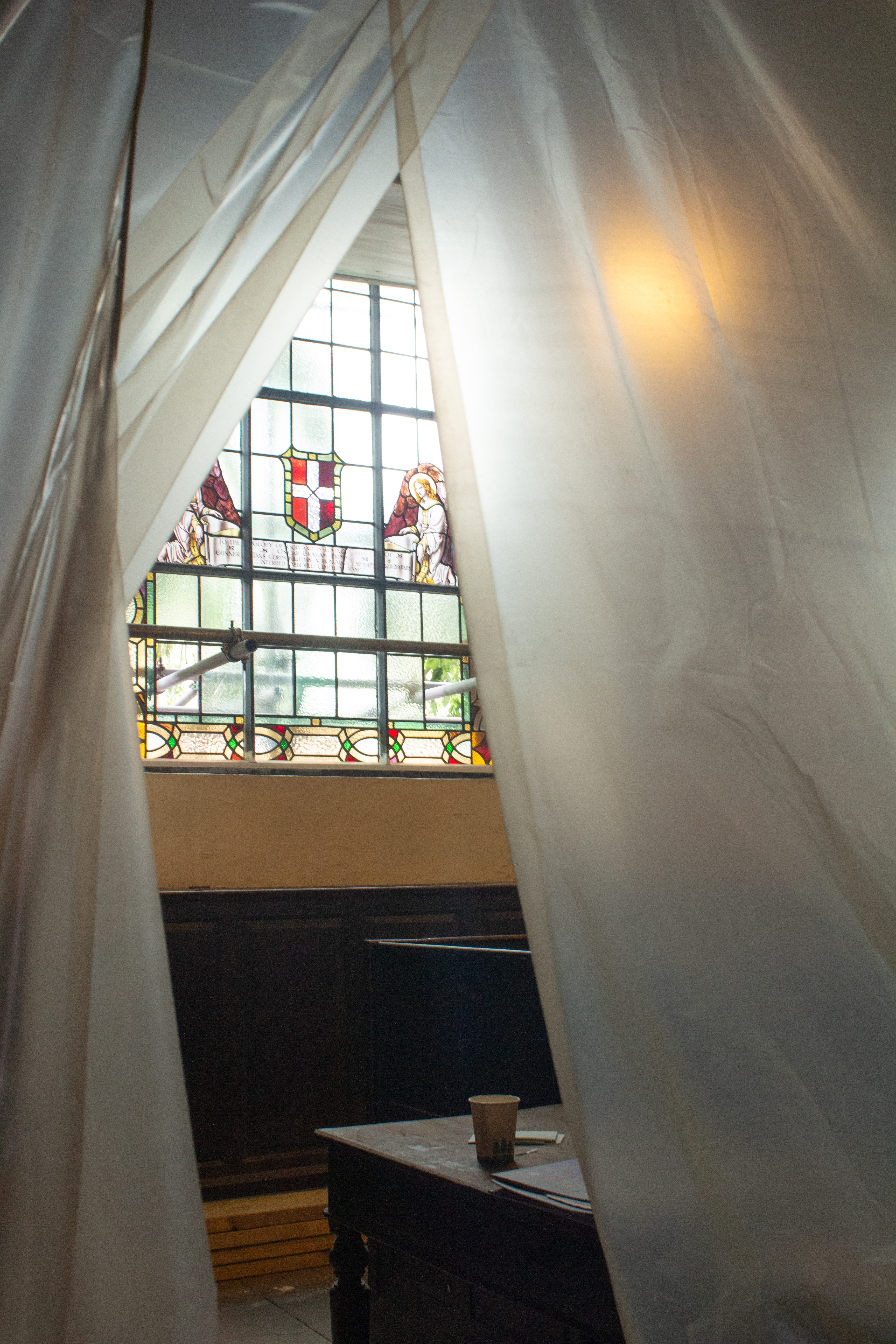 St. Johns Lancaster interior showing a window with heraldry seen through a gap in protective tarp like a curtain. A paper cup rests on a pew.