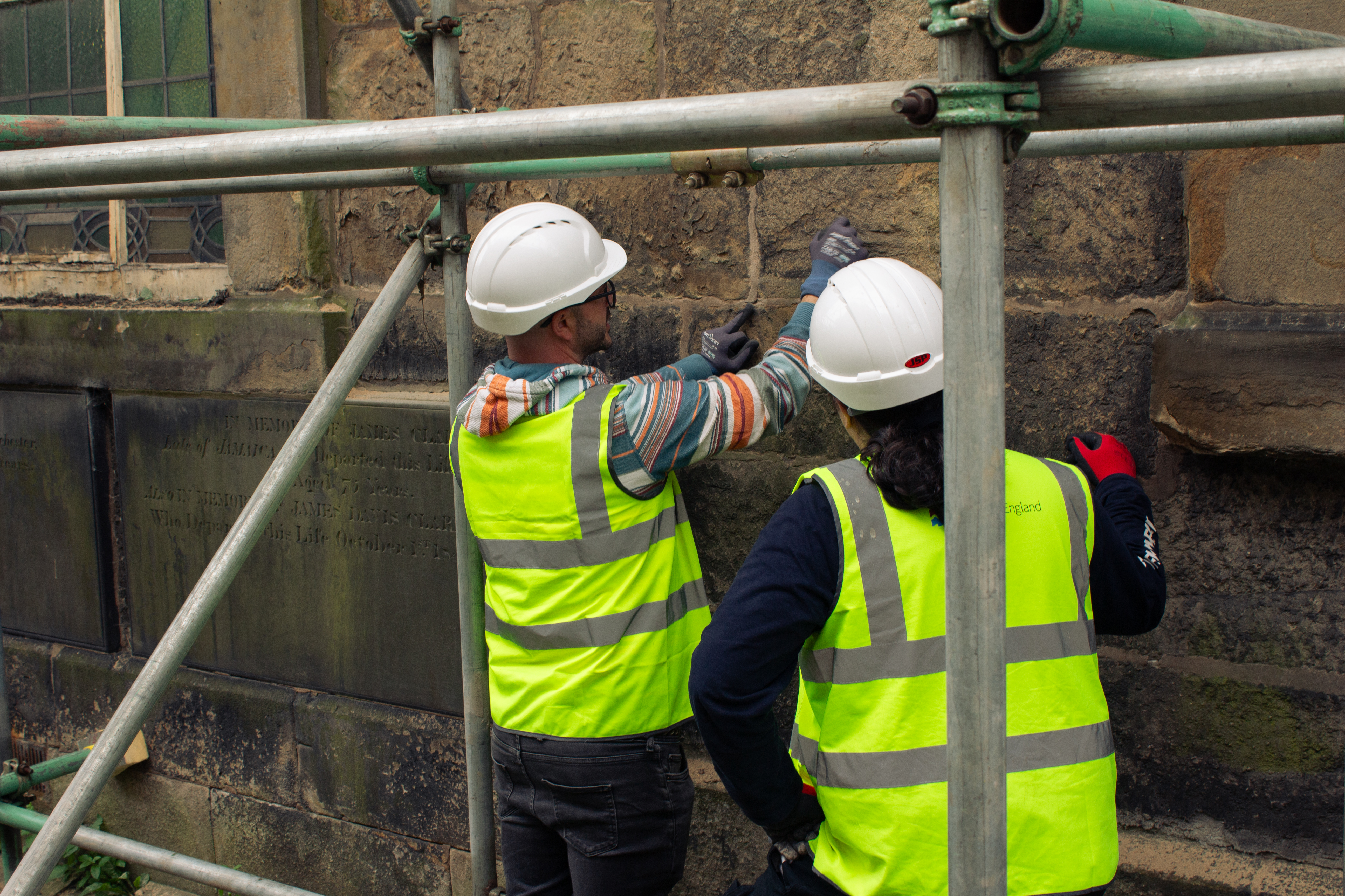 A master crafts person explaining replacing lime mortar to an apprentice indicating old mortar on a wall at St. Johns Lancaster.